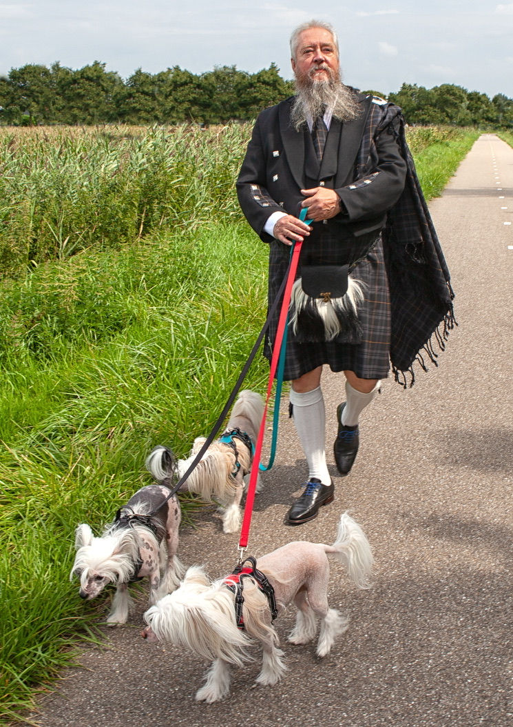 Job in Schotse kleding, wandelend met drie honden op een pad langs het groen.